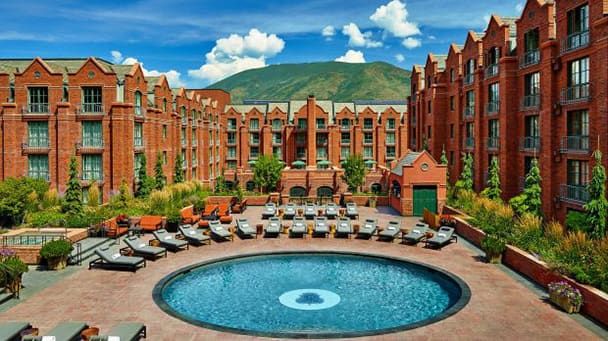 A circular outdoor pool surrounded by lounge chairs sits in the courtyard of a large red-brick hotel with mountain views in the background under a partly cloudy sky.