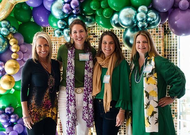Four women stand smiling in front of a backdrop decorated with green, purple, and gold balloons, wearing casual clothes and scarves.