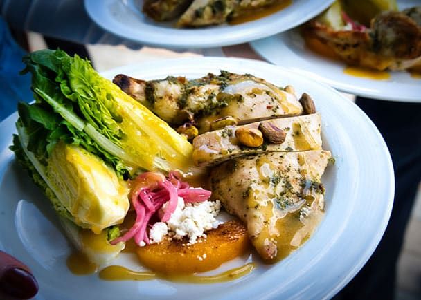 A plate with sliced grilled chicken, romaine lettuce, pickled onions, feta cheese, pistachios, and a golden sauce, served with a slice of roasted squash.