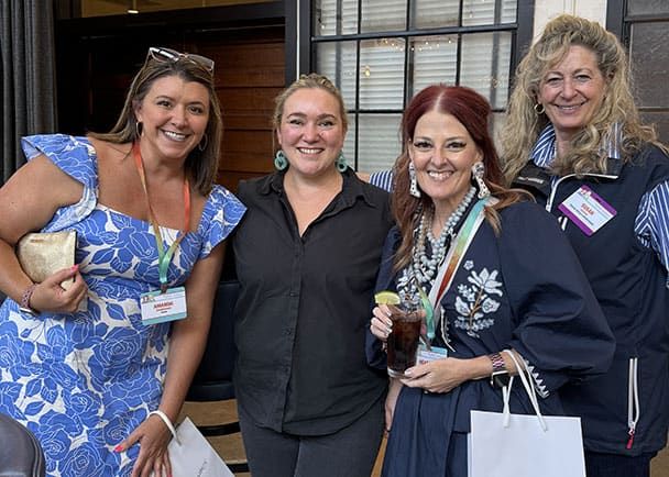 Four women pose together indoors, wearing conference name badges. One woman holds a drink and another carries a clutch. They are smiling at the camera.