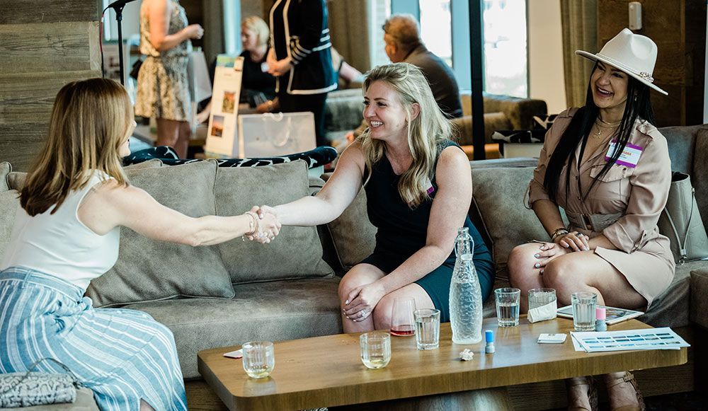 Three women sitting on sofas in a lounge area. One, an event planner, is shaking hands with another, while a third woman looks on, smiling. Several glasses and items are placed on the table in front of them.