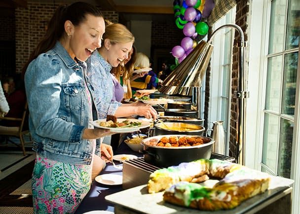 Two happy, smiling women serve themselves food from a buffet table indoors, with colorful balloons and sunlight streaming through nearby windows.