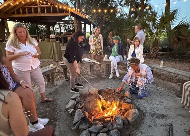 A group of people sit and stand around a campfire, roasting marshmallows, with a wooden pavilion and string lights in the background.