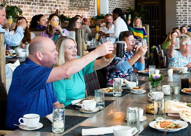 A group of people sitting around a large table in a restaurant, raising their glasses in a toast. Plates, glasses, and cups are visible on the table.