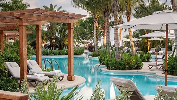 Rosewood Baha Mar resort pool area with lounge chairs, umbrellas, wooden pergolas, and lush tropical plants surrounding a curved swimming pool.