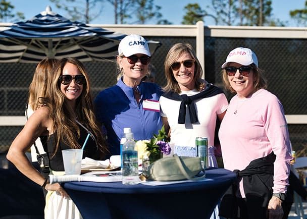 Four women stand together smiling outdoors by a table with drinks and flowers, wearing casual clothing, sunglasses, and hats. A fence and trees are visible in the background.