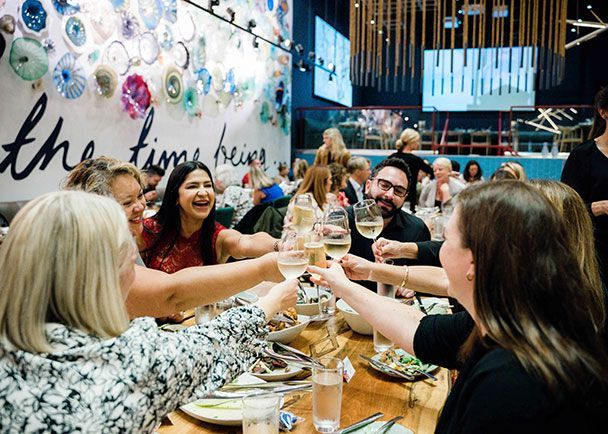 A group of people sitting around a restaurant table raise their glasses in a toast, smiling and enjoying a meal together.