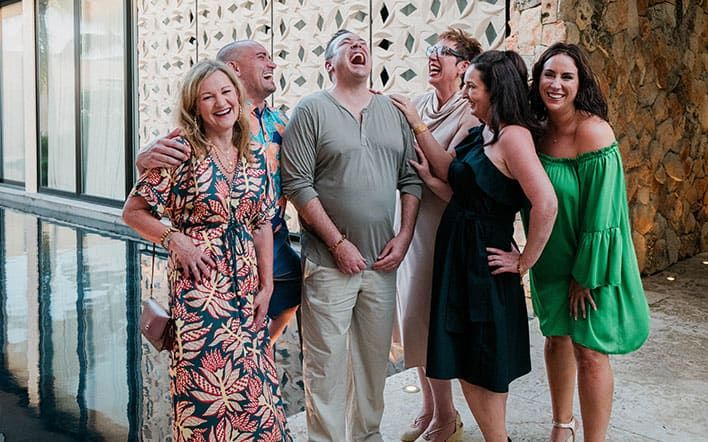 A group of six adults stand together laughing and smiling near a reflective pool, with a stone wall and modern building in the background.