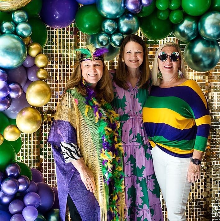 Three women pose in front of a gold sequin backdrop with green, purple, and gold balloons; one wears Mardi Gras attire, another a floral dress, and the third a striped shirt and sunglasses.