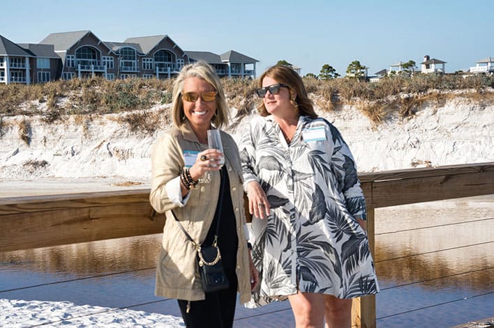 Two women stand on a wooden boardwalk at the beach, wearing sunglasses and casual clothing, with houses and sand dunes in the background.