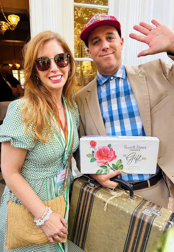 A woman in a green dress and a man in a beige suit hold a suitcase and a Russell Stover candy box while standing outside and smiling at the camera.
