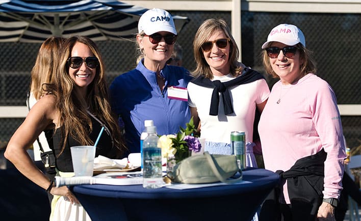 Four women wearing sunglasses and casual clothing stand around a cocktail table outdoors, smiling at the camera.