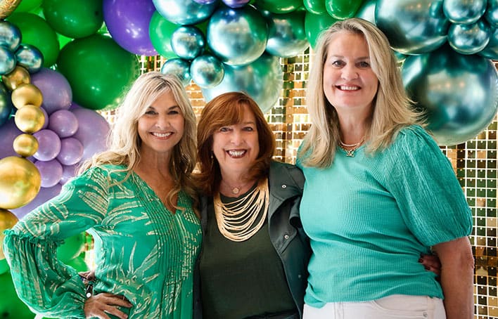 Three women standing and smiling in front of a decorative balloon backdrop featuring green, purple, and gold balloons.