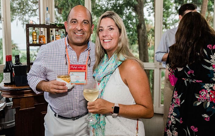 A man and woman stand indoors at a social event, smiling at the camera. The man holds a plate of food, and the woman holds a glass of white wine. Shelves and trees are visible in the background.