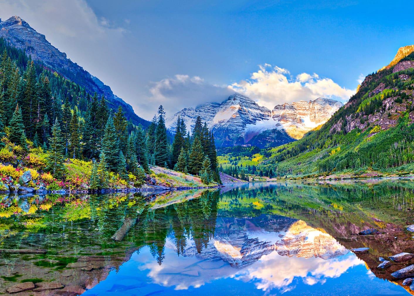 Maroon Bells: A clear lake reflects pine trees and snow-capped mountains under a bright blue sky with scattered clouds in a scenic valley.