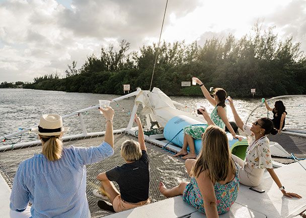 A group of people sit and stand on the deck of a boat, raising drinks and looking toward the water and treeline under a partly cloudy sky.