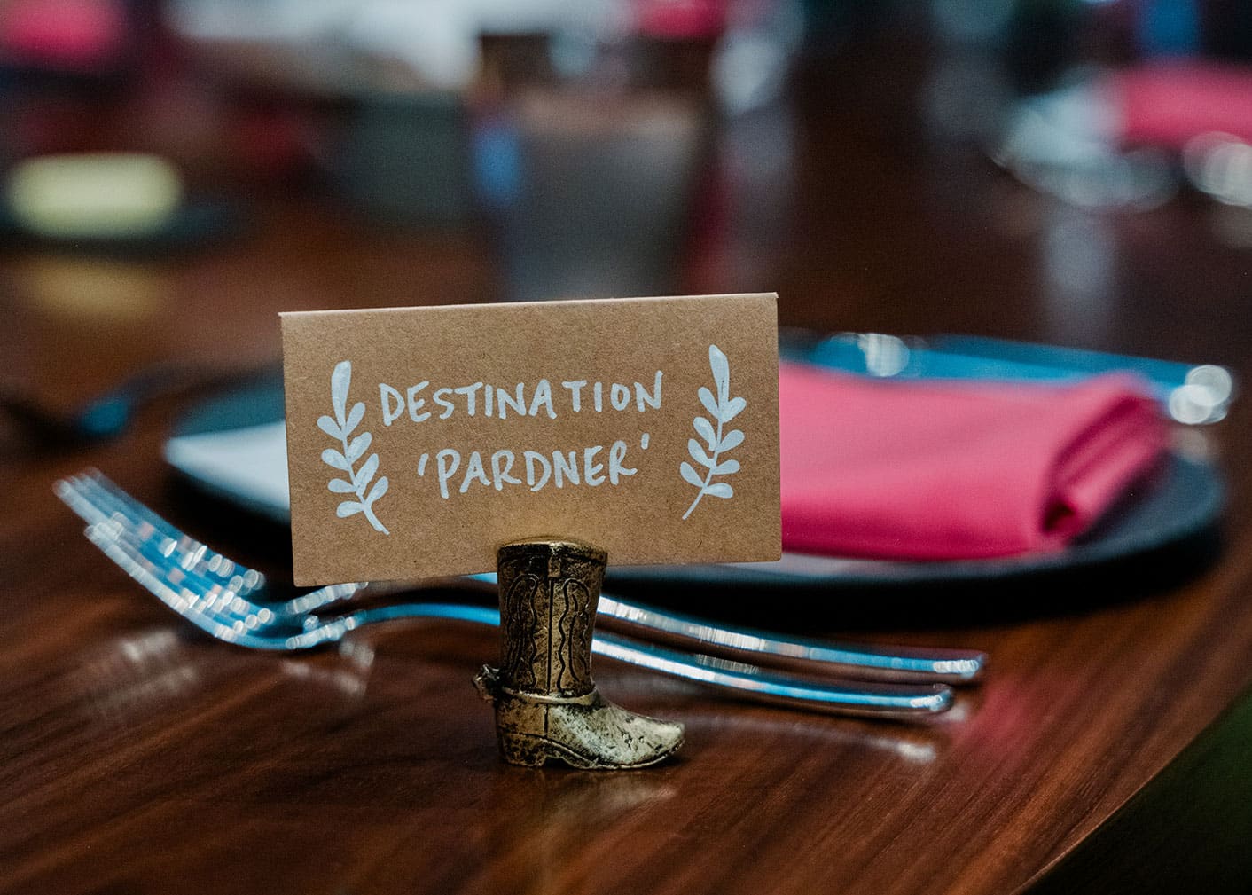 A small card reading "Destination 'Pardner'" is held by a miniature cowboy boot stand on a wooden table set with silverware and pink napkins.