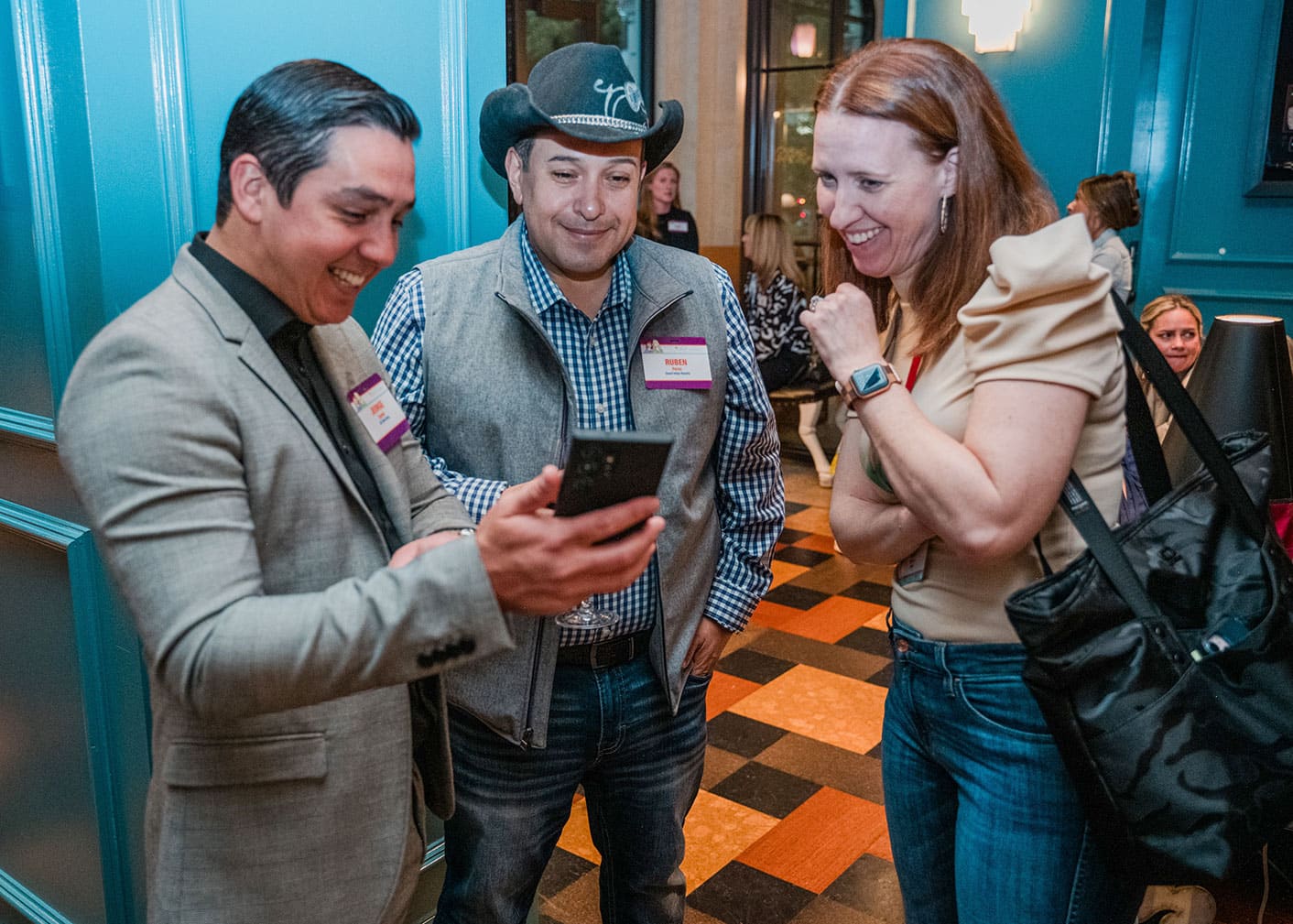 Three people stand indoors looking at a smartphone, smiling and engaged in conversation; two wear name tags, and one wears a cowboy hat.