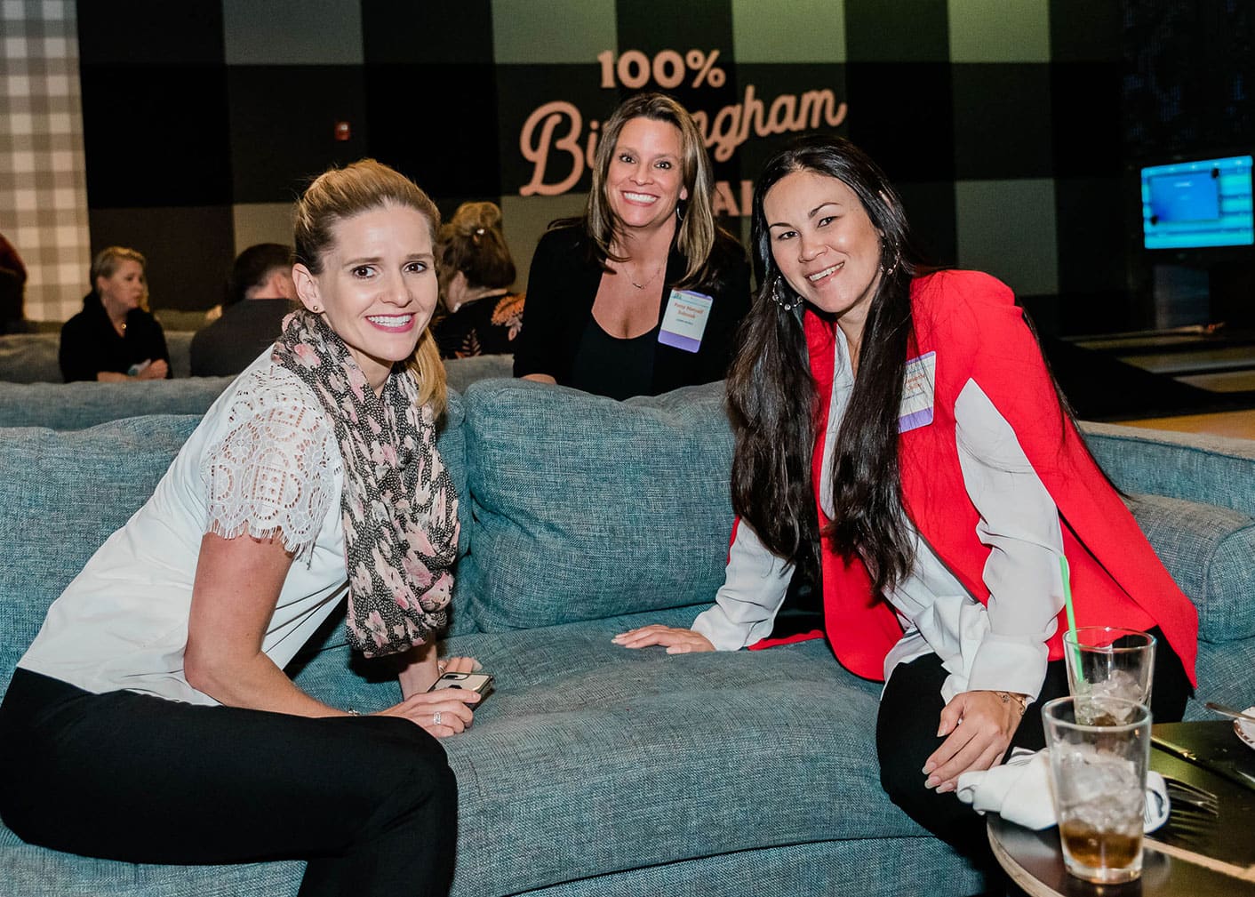 Three women sit and smile on a blue couch at an indoor event; drinks are on the table in front of them, and a sign in the background reads &ldquo;100% Birmingham&rdquo;.