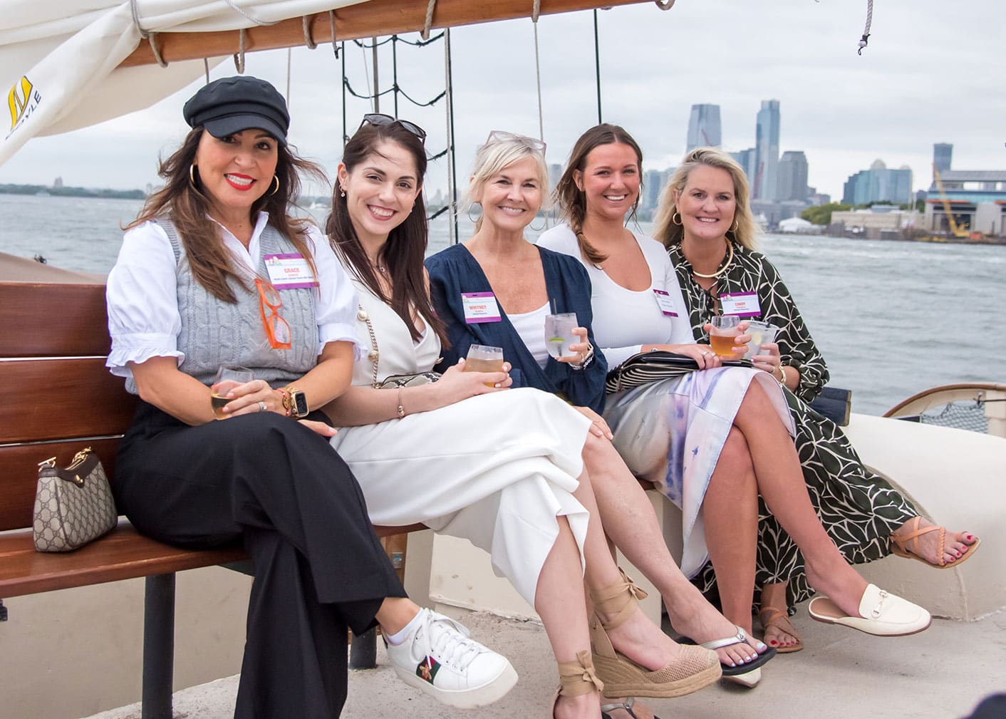 Five women sit on a bench on a boat, smiling and holding drinks, with city buildings and water visible in the background.
