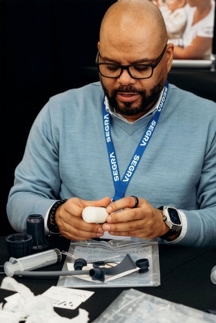 A man wearing glasses and a blue lanyard sits at a table assembling a water filter, with various components and tools spread out in front of him.