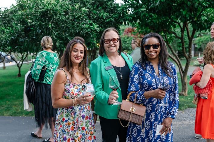 Three women stand outdoors holding drinks, smiling at the camera, with trees and other people in the background.