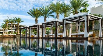 Chileno Bay Resort view of a row of poolside cabanas with white curtains and cushioned seating, surrounded by palm trees, reflected in a still swimming pool under a partly cloudy sky.