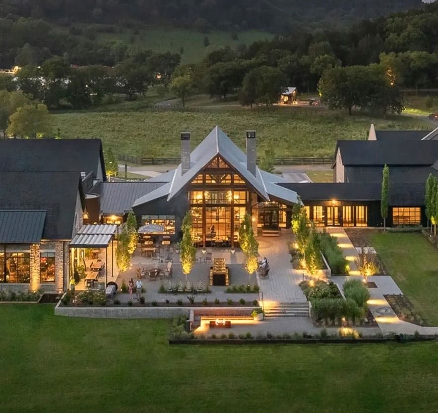 Southall Farm & Inn. View of a large modern building with glass walls, outdoor seating areas, and landscaped gardens, illuminated by lights in a rural, green setting at dusk.