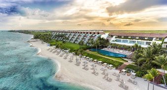 Aerial view of a beachfront Grand Velas Riviera Maya resort with rows of lounge chairs and umbrellas along the sandy shore, a pool, and multiple buildings surrounded by palm trees at sunset.