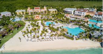 Aerial view of Sanctuary Cap Cana beachfront resort with pools, palm trees, lounge chairs, and several large buildings next to the ocean.