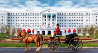 A horse-drawn carriage with a driver in a red coat is in front of the large white Greenbrier Resort hotel building with red awnings and flags.