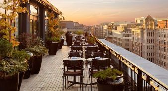 Rooftop restaurant at Riggs DC in Washington, DC, with tables and chairs set for dining, surrounded by potted plants, overlooking city buildings at sunset.