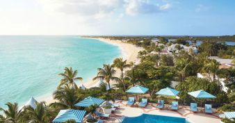 A coastal resort with a swimming pool, blue and white umbrellas, palm trees, and a sandy beach next to clear turquoise sea under a partly cloudy sky.