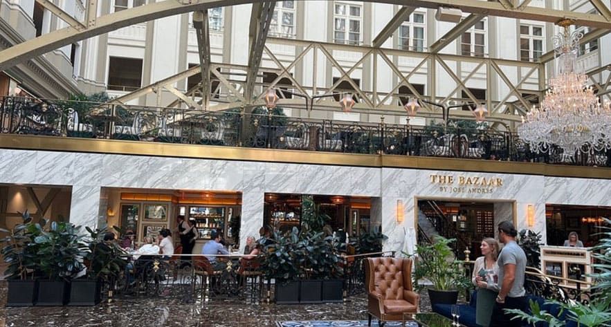 People sit and stand in a spacious indoor atrium with marble walls, a chandelier, and a restaurant named "The Bazaar by José Andrés." The ceiling has large metal beams and windows.