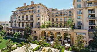The Maybourne Beverly Hills - a multi-story luxury hotel with balconies, arched windows, and outdoor seating areas surrounded by manicured gardens and palm trees.