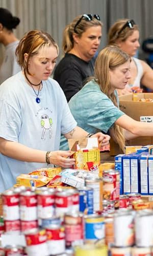 Several people sort and organize canned goods and boxed food items on a table, volunteering with Wine To Water.
