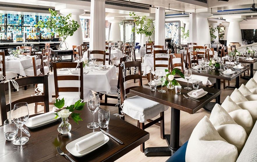 Spacious KYMA restaurant dining area with wooden tables and chairs, white tablecloths, neatly set glassware, and green plants as decor.