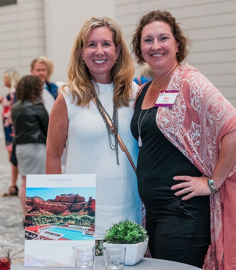 Two women standing and smiling at aDuring a Retreats Resources Roadshow event, with a display of a The Enchantment Resort photo and small plants on the table in front of them.