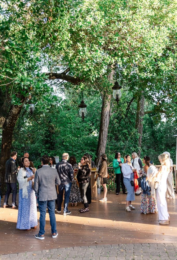 A group of people gather and converse on a wooden deck outdoors, surrounded by tall trees and hanging lanterns in daylight.