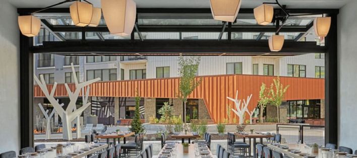 KITA Restaurant at Pendry Park City Resort - A modern restaurant dining area with long tables and chairs, open to an outdoor patio with contemporary decor and geometric white sculptures, facing a building with orange and beige panels.