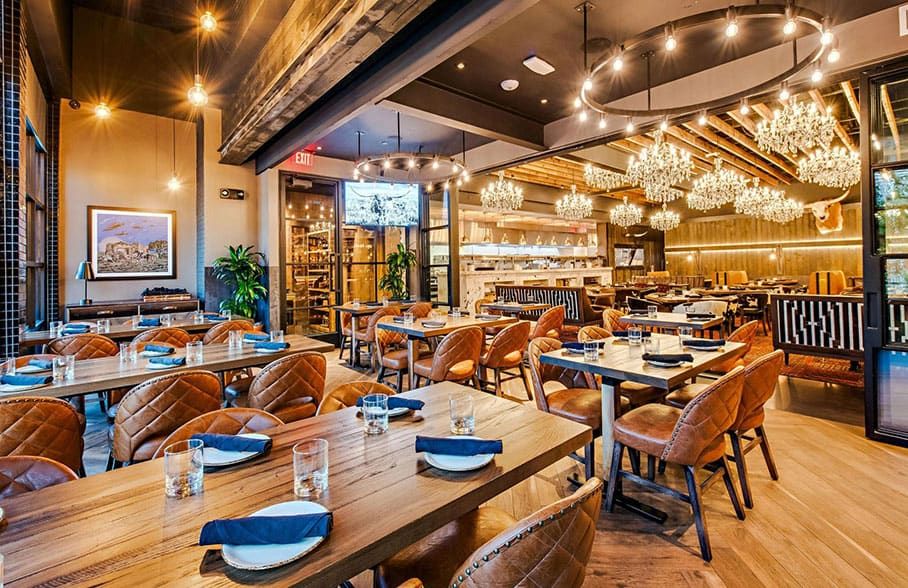 Haywire, Austin, TX – A modern restaurant dining area with wooden tables, brown leather chairs, glassware, folded napkins, chandeliers, and an open kitchen in the background.