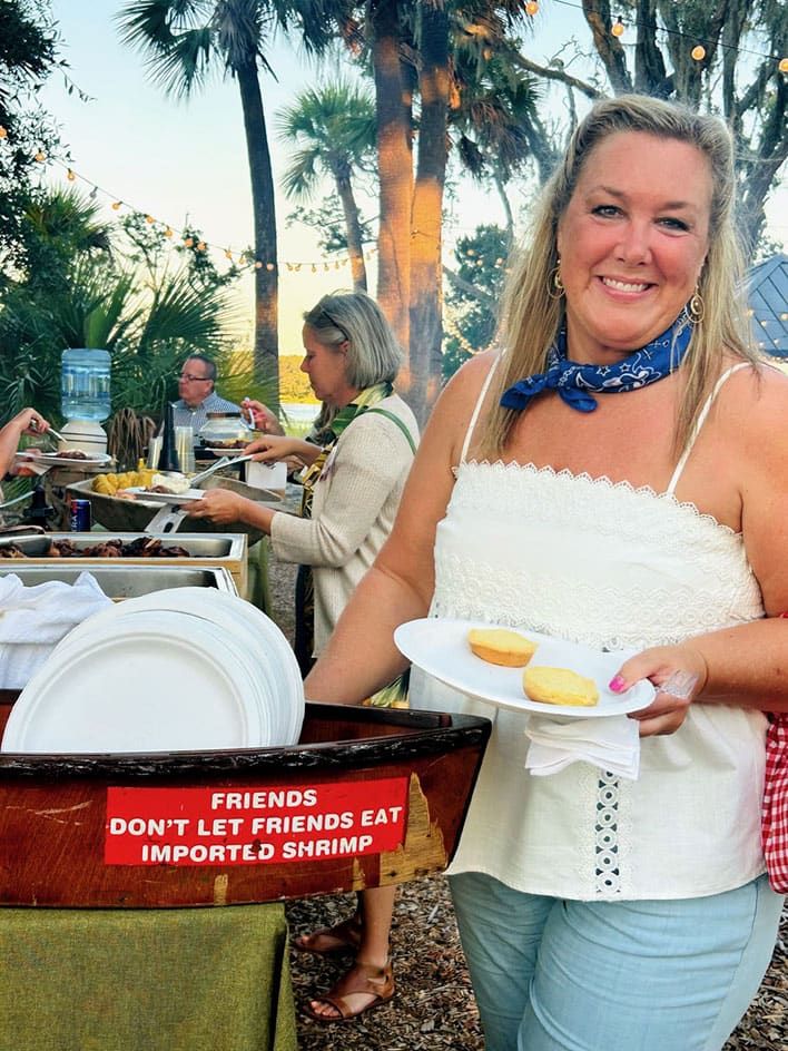 A woman holding a plate with two biscuits stands by a food serving table outdoors. A sign on the table reads: “Friends don’t let friends eat imported shrimp.”