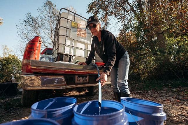 A woman fills blue plastic buckets with water from a large tank in the back of a pickup truck outdoors on a sunny day.