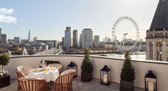 Corinthia Hotels royal penthouse rooftop. Table with breakfast setup on a rooftop terrace overlooking London landmarks, including the London Eye, the Shard, and city buildings under a clear sky.