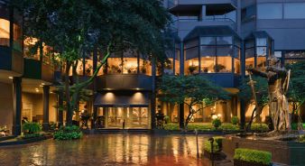 Entrance of the modern Windsor Court New Orleans hotel building at dusk, with large windows lit from inside, trees, landscaped greenery, and a statue fountain in the foreground.