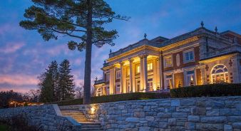 Canyon Ranch - Tucson, AZ. Large neoclassical mansion with columns and exterior lighting, set behind a stone wall and stairs, surrounded by trees at dusk.