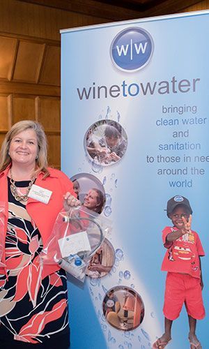 At a Wine To Water filter build, a woman holding a clear bag stands beside a promotional banner promoting clean water and sanitation for those in need globally.