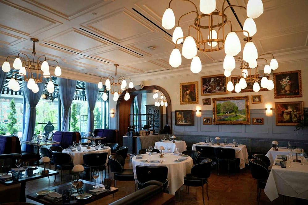 Chardon, Houston, TX – Elegant restaurant interior with round tables covered in white cloths, modern black chairs, large light fixtures, blue curtains, and framed artwork on the walls.