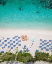 Aerial view of a sandy beach with rows of blue lounge chairs, kayaks arranged in the center, and clear turquoise water bordering the shore.