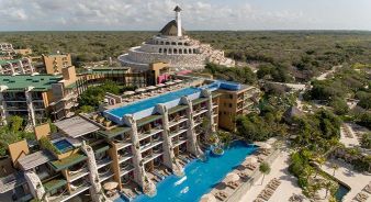 Aerial view of Hotel Xcaret Mexico with tiered pools, lounge chairs, and a unique dome-shaped building surrounded by greenery under a partly cloudy sky.
